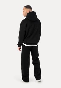 Mann steht und ist leicht nach links gedreht, trägt einen schwarzen Hoodie, schwarze Hose, weiße Sneakers und einen sichtbaren weißen Hemdsaum.