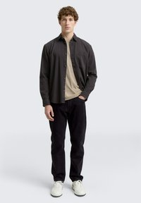Ungdommelig mann som står med en beige skjorte under en åpen svart skjorte med knapper, svarte bukser og hvite sneakers på en ensfarget bakgrunn.