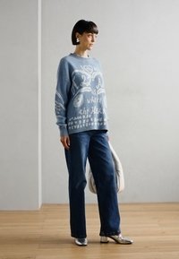 Lyseblå strikkegenser med hvit tekst og mønster, kombinert med mørkeblå jeans og sølvfargede sko. Modellen holder en stor hvit veske.