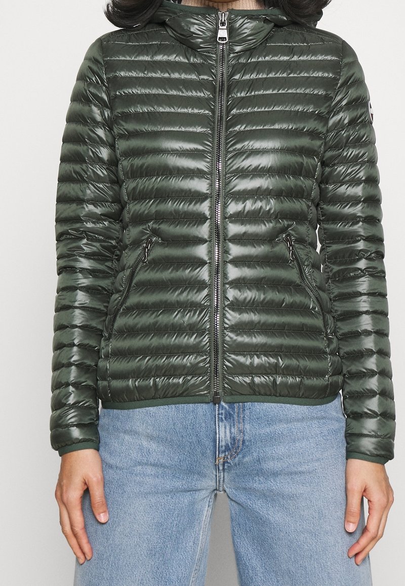 Zelená péřová bunda se lesklou texturou, vertikálním prošíváním, předním zipem, bočními kapsami a kapucí. Nositá přes světle modré džíny.
