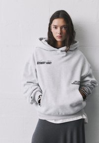 Sivá mikina s veľkou prednou kapsou, kapucňou a rebrovanými manžetami. Na prednej strane a na spodnej časti má potlačený text "STREET UNIT" a ďalšie grafiky.