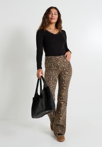Haut noir à manches longues avec bordure en dentelle, associé à un pantalon évasé à imprimé léopard. Le modèle tient un sac fourre-tout noir. Chaussures marron avec un motif.
