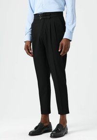 Pantalon noir fuselé avec un devant plissé, doté d'une fermeture à double boucle à la taille et d'un tissu lisse, associé à des mocassins noirs.