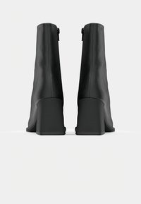 Botas negras hasta el tobillo con un acabado de cuero suave, tacones altos de bloque y un detalle de cremallera trasera para facilitar su uso. Diseño estructurado y en forma cónica.
