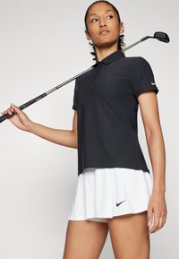 Black polo shirt made of lightweight fabric, paired with a white skirt. Skirt features a black Nike logo. Golf club held in one hand.