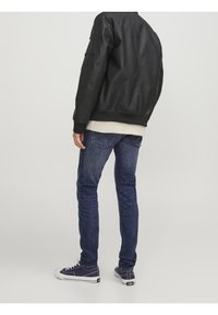 Giubbotto bomber in pelle nera con polsini e cintura a coste, abbinato a jeans in denim blu. Sneakers bianche e blu navy completano il look.