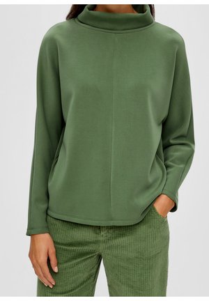 Femme portant un haut vert olive à manches longues et col montant ainsi qu'un pantalon côtelé assorti, une main dans la poche.