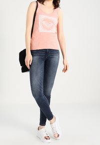 Top sin mangas de color rosa claro con un diseño gráfico blanco, combinado con jeans ajustados azul oscuro y zapatillas blancas con acentos metálicos. Bolso negro.