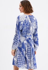 Vestido de estampado azul y blanco con mangas largas, cintura ajustada y falda fluida. Presenta diseños florales y geométricos. Textura de tela suave.