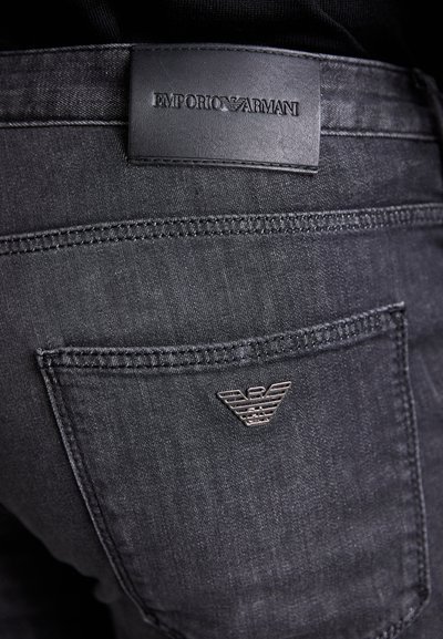 Crne traperice od denima s kožnom oznakom Emporio Armani na pojasu i malim metalnim logom iznad stražnjeg džepa.