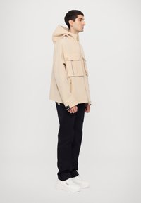 Beige hooded jacket with a zip closure and front pockets, paired with black pants and white sneakers, displayed on a neutral background.