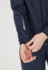 Dunkelblaues Sportjackett mit glatter Textur, ausgestattet mit einem Reißverschluss und reflektierenden Streifen an den Ärmeln. Hände, die den Bund anpassen.