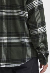 Cămașă din flanel în carouri verzi și negre cu dungi albe, având manșete cu nasturi și un material moale, texturat. Croiala este ușor curbată.