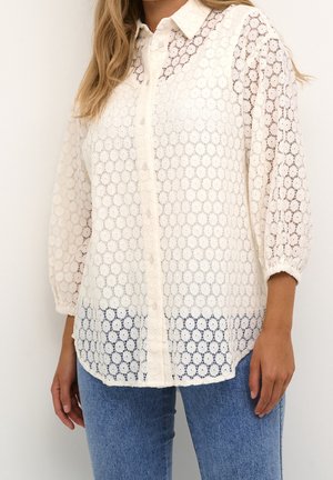 Femme portant un chemisier en dentelle blanc boutonné avec des manches bouffantes 3/4, associé à un jean bleu, visible du cou jusqu'aux hanches.