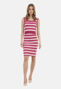 Abito aderente senza maniche con un motivo a zigzag rosa, bianco e lavanda. Tessuto a maglia con una cintura di colore uniforme. Indossato con tacchi alti color nude.