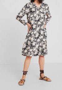 Sukienka z długim rękawem w czarno-kremowy kwiatowy wzór, z dekoltem w serek i bocznymi kieszeniami, zestawiona z brązowymi sandałami na cienkich paskach. Gładka faktura materiału.