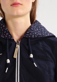 Close-up of a person wearing a navy zip-up jacket with floral-patterned hood lining and navy drawstrings with white beads.