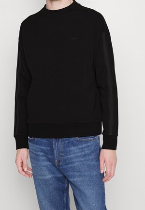 Sudadera negra de manga larga, con escote redondo y puños a juego. Presenta un sutil logo en los laterales. Combinada con jeans de mezclilla azul.