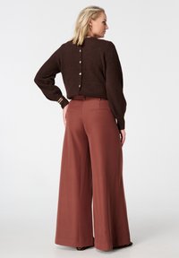 Bruine trui met knoopdetail aan de achterkant, gecombineerd met broek in roestkleur met wijde pijpen. Zachte textuur, comfortabele pasvorm en gladde stof.