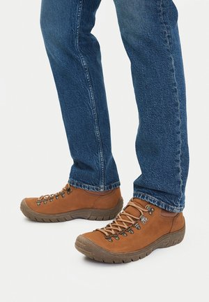 Brązowe buty trekkingowe z gumowymi podeszwami, metalowymi oczkami i detalami z sznurowadeł, zestawione z niebieskimi dżinsami o delikatnym wyblakłym efekcie i prostym kroju.