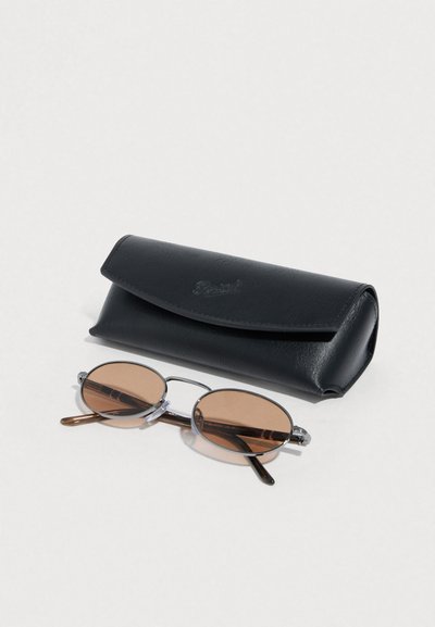 Οβάλ γυαλιά ηλίου με καφέ φακούς και διάφανους σκελετούς δίπλα σε μαύρη δερμάτινη θήκη. Η θήκη διαθέτει κλείσιμο με καπάκι και ανάγλυφο λογότυπο.