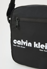 Svart syntetisk skulderveske med glidelås, med en jevn tekstur og fet hvit skrift som viser "calvin klein" og stedsdetaljer.