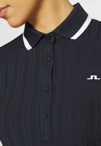 Svart pikétröja med ribbad textur, knäppning framtill och kontrasterande vit krage. Logotyp i vitt på bröstet.