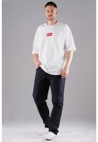 White short-sleeve t-shirt with a red logo patch, paired with dark navy pants and white sneakers. Simple design with relaxed fit.