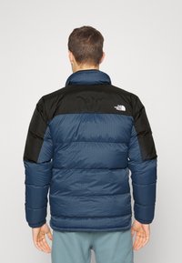Puffer jacket in navy blue with black accents, featuring a high collar, quilted design, and ribbed cuffs. Logo on the upper back.