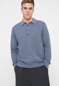 Blauer Langarm-Polo-Pullover, strukturierter Strick, mit Kragen und drei schwarzen Knöpfen an der Vorderseite. Kombiniert mit dunklen Hosen.