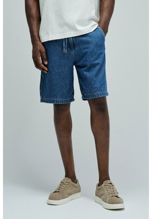 Hombre con pantalones vaqueros cortos azules de longitud media, camiseta blanca y zapatillas beige, de pie con una mano en el bolsillo sobre fondo liso.