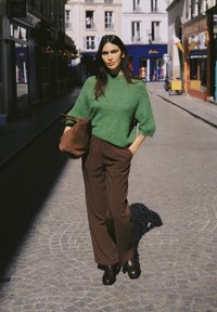 Femme aux longs cheveux foncés portant un pull vert et un pantalon marron, portant un sac marron, debout dans une rue pavée ensoleillée.