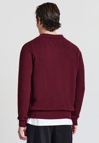 Maglioncino bordeaux a costine con colletto polo, maniche lunghe e tessuto strutturato. Vestibilità leggermente aderente, indossato sopra una camicia bianca.