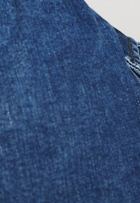 Lähikuva sinisestä denim-kankaasta, jossa näkyy saumoja ja pintarakennetta, ja oikealla puolella näkyy osa taskusta.