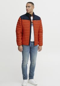 Polstret jakke i orange og marineblå med høj krave, lynlås foran og quiltet tekstur. Sammen med lyseblå jeans og hvide sneakers.