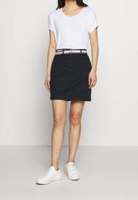 Zwarte denim rok met rechte snit, voorzien van een tailleband met een riem met meerkleurige strepen. Gecombineerd met een wit T-shirt en witte sneakers.