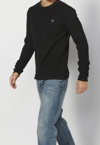 Černé vy ribbené tričko s dlouhým rukávem, kulatým výstřihem a malým logem na hrudi, spárované s světle modrými džínami.