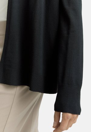 Hand met een zwart lange mouwen trui met geribbelde manchetten, rustend nabij beige broek tegen een effen lichte achtergrond.