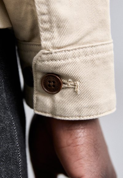 Manga de chaqueta de mezclilla de algodón beige con un botón marrón de cierre, que presenta una textura acanalada diagonal y detalles de costura visibles.