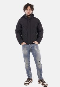 Uomo con capelli ricci che indossa una giacca nera con cappuccio, jeans chiari strappati e sneakers marroni e bianche, in piedi con le mani nelle tasche.