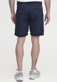 Pantalones cortos azul marino de algodón con dobladillos enrollados, dos bolsillos traseros con botones marrones, usados con zapatillas grises y un calcetín blanco.
