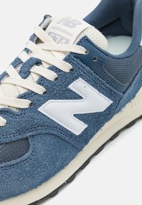 Kék velúr sneaker hálós betétekkel, krémszínű fűzőkkel és fehér logóval. Jellemzője a struktúrált felsőrész és egy fekete gumitalp.