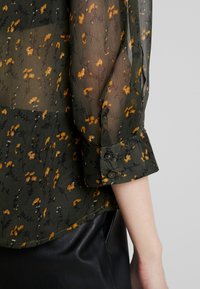 Hihna kukallinen paita tummanvihreänä, jossa on oransseja kukkia, pitkät hihat ja napilliset hihansuut. Sileä rakenne kerroksellisella suunnittelulla.