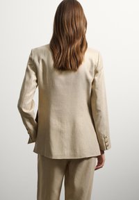 Blazer de lino beige con mangas largas, hombros estructurados y detalles de botones dorados, combinado con pantalones a juego, vista desde la parte trasera.