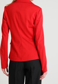 Blazer ajusté rouge avec col cranté, manches longues et coutures structurées, associé à un pantalon noir, texture de tissu lisse.