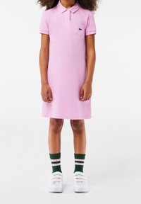 Růžové polo šaty s krátkými rukávy, límečkem a malou náprsní kapsou. Spojené se zeleno-růžovými pruhovanými ponožkami a bílými botami.