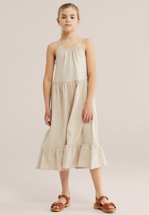 Ragazza giovane in piedi che indossa un vestito midi beige senza maniche e sandali marroni con cinturino e fibbia su uno sfondo semplice.