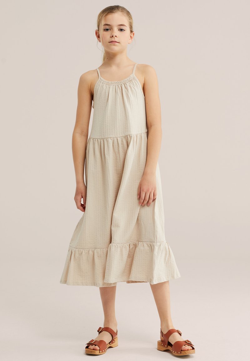 Jeune fille debout portant une robe midi beige sans manches et des sandales marron à brides avec boucle sur un fond uni.