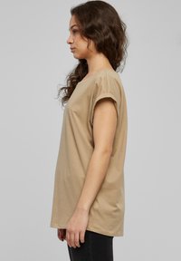 Beige kortärmad T-shirt i mjukt bomullsmaterial, med lös passform, rund halsringning och rullade ärmar, samt subtila sömdetaljer.