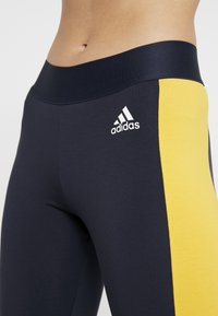 Granatowe legginsy z żółtym panelem bocznym, wykonane z elastycznego materiału. Posiadają solidny czarny pas w talii i białe logo Adidas.